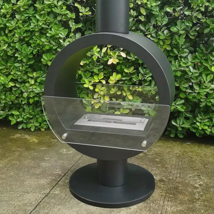 bioethanol-haengekamine-zenica-decke-ethanolkamin-schwarz-deckenkamin-deckenmontage-mit-glas-mit-flammen-rund-mit-standfuss-aussenbereich-vor-hecke-auf-betonboden